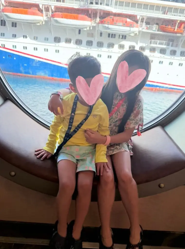Two kids sitting in a Disney cruise ship porthole window wearing lanyards with Key to the World cards, cruise ship visible outside.