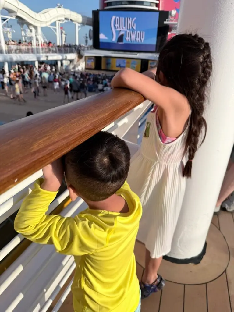 Two young kids from behind leaning on a Disney cruise ship railing watching the Sailing Away deck party on embarkation day.