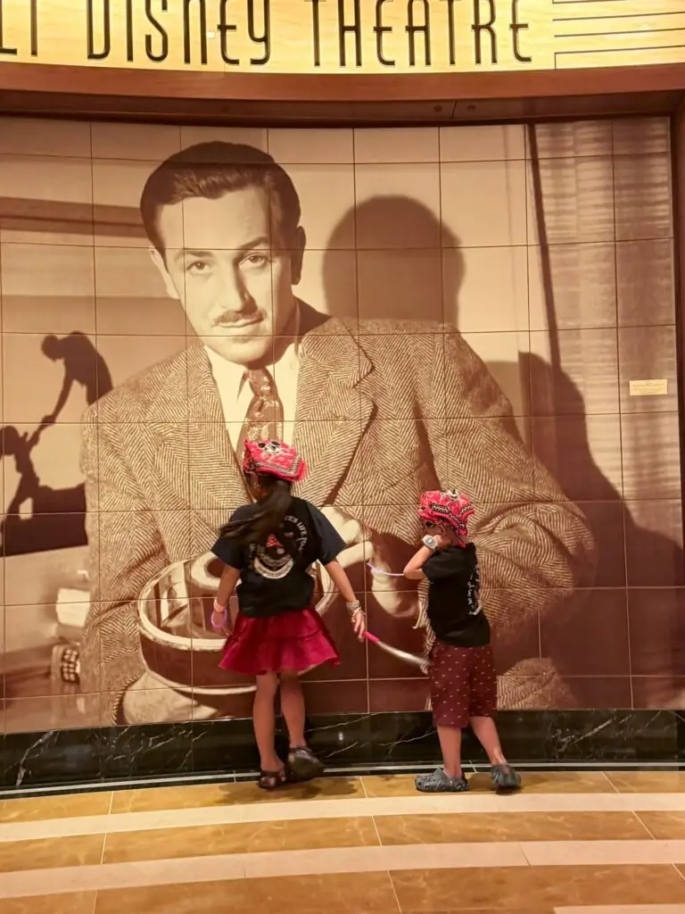 Two kids from behind in pirate night gear including pink pirate hats and black shirts standing in front of the Walt Disney Theatre mural on the Disney Fantasy.